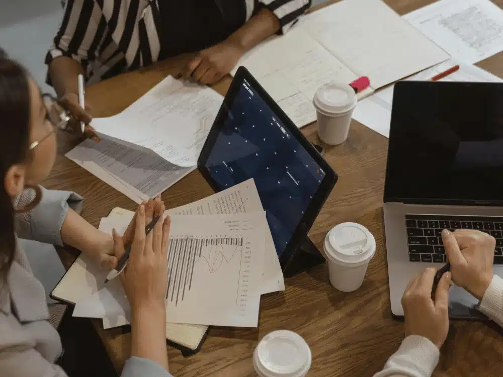 Team discussion: professionals with coffee cups, laptops, and data sheets.