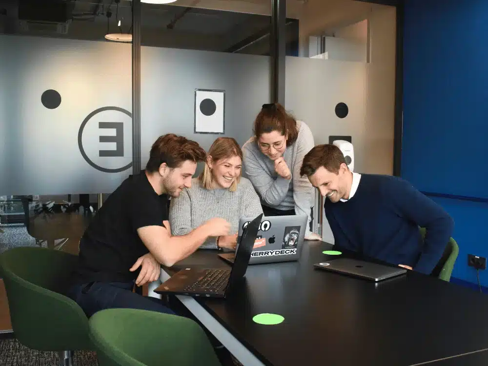 Enthusiastic professionals discussing work, with a laptop open on the desk in a modern office setting.