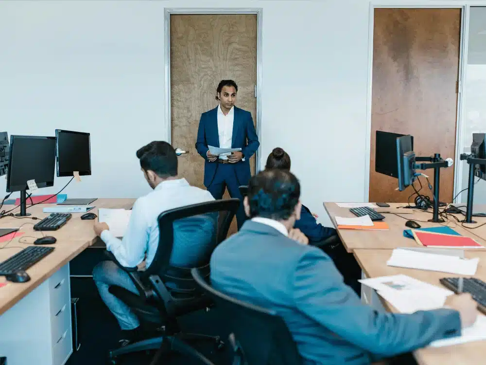 Man in a blue suit presenting to coworkers during a business meeting in an office.
