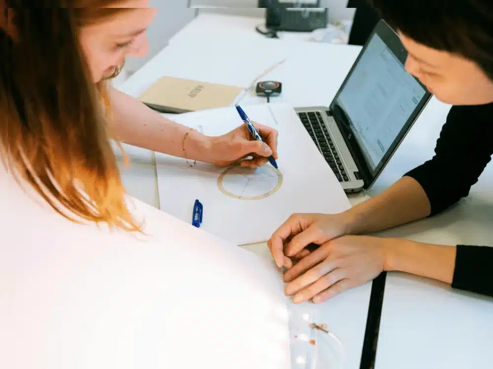 Two hands engage in a collaborative design process, drawing on paper with a blue pen, while a laptop displays design software in the background.